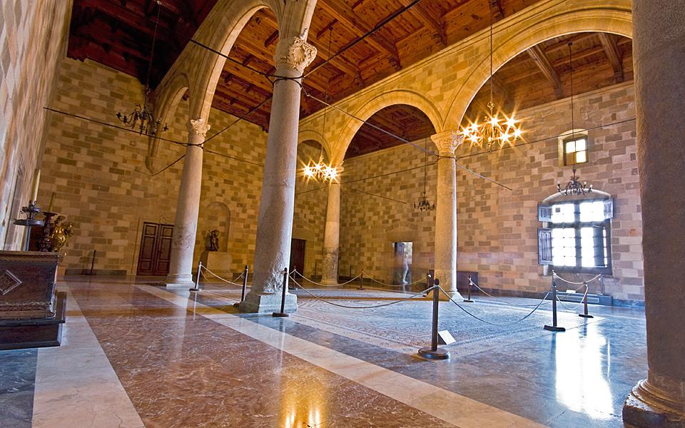 Interior of the Palace of the Grand Master — a vast stone hall with marble floors, columns, chandeliers and a timber-beamed ceiling