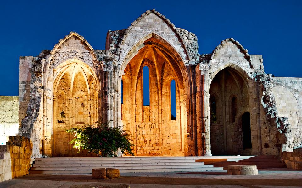 Illuminated Gothic ruins in Rhodes Old Town at dusk — three arched stone facades lit warmly against a deep blue evening sky