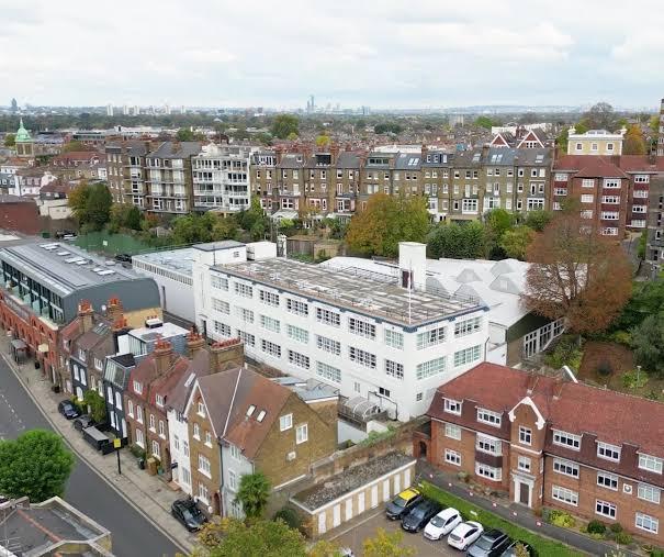 An aerial view of the Poppy Factory building in Richmond — a white, low-rise industrial building with a distinctive sawtooth roof, tucked among Victorian mansion blocks, brick houses and mature trees, with the London skyline in the distance