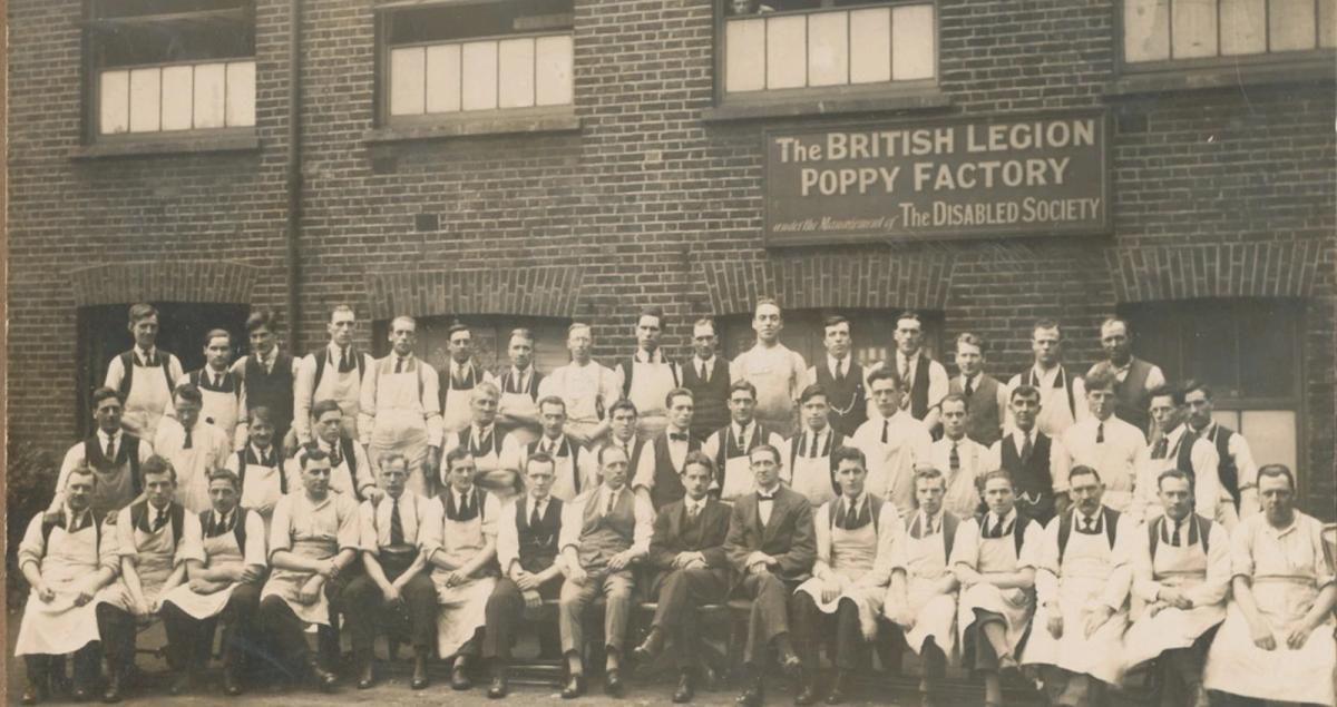A sepia photograph of Poppy Factory workers in aprons and ties gathered outside the original British Legion Poppy Factory building, under a sign reading 'The British Legion Poppy Factory under the Management of The Disabled Society'