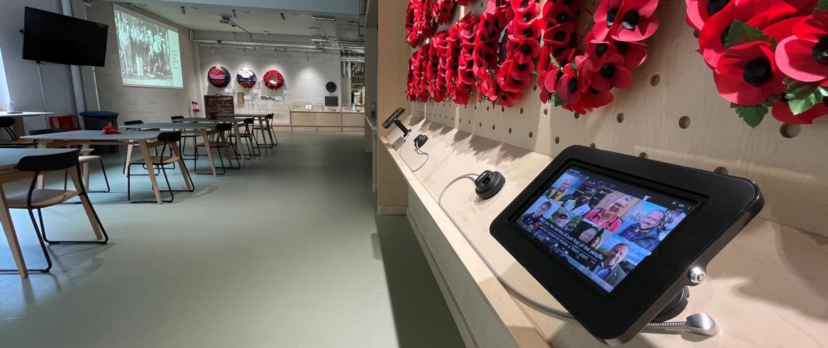 The Poppy Factory visitor centre in Richmond — a bright, modern space with a wall of hand-made red Remembrance wreaths and poppies on the right, an interactive tablet in the foreground, and long tables, wreaths on display and a projected archive image at the far end