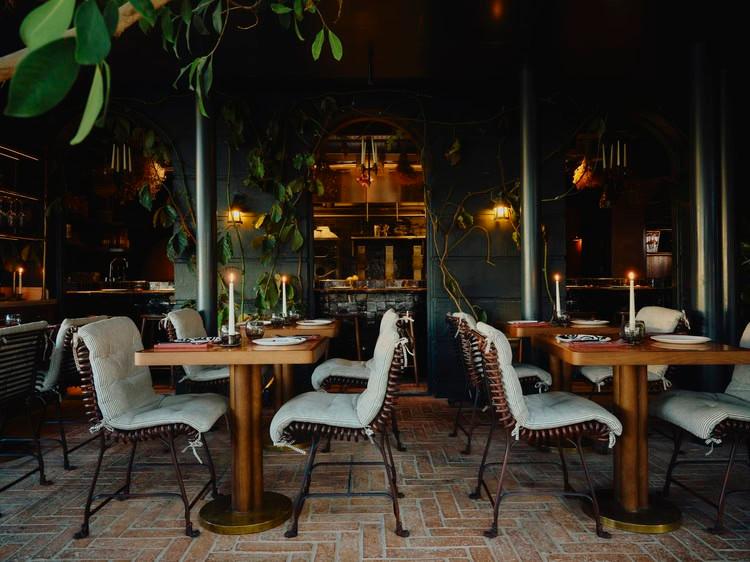 Tower House, Richmond — the candlelit main dining room with dark walls, iron pillars trailing with ivy and vines, brick floor, and wooden tables set with linen cushions and taper candles