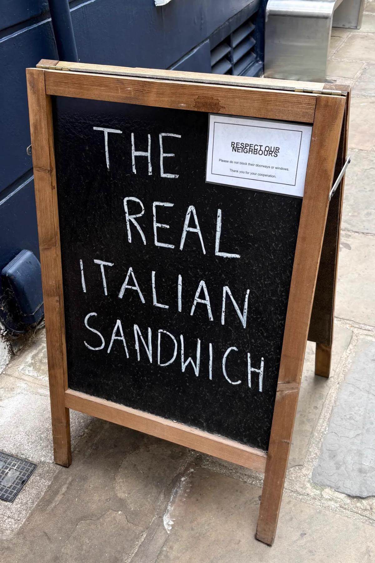 Chalkboard A-board outside Via Romana Deli, Richmond, reading 'The Real Italian Sandwich' — the shop's one-line proposition